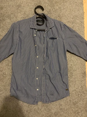 Randig skjorta från Scotch & Soda - Blå och vit randig skjorta från Scotch & Soda i storlek S. Skjortan har klassisk krage, knäppning framtill och en bröstficka med liten detalj. Tillverkad i bomull och passar perfekt för en stilren look.