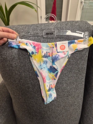 Färgglad bikiniunderdel från Bik Bok - Säljer en bikiniunderdel från Bik Bok i storlek XS med högt skuren modell. Tyget har ett färgstarkt abstrakt mönster i blått, rosa, gult och vitt. Materialet är mjuk polyester med elastan som ger bra stretch och passform. Perfekt för strandhäng och poolparty!