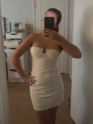 Vit axelbandslös kortklänning - Säljer en snygg vit kortklänning med markerad byst och axelbandslös design. Klänningen har figurnära passform och sömmar som framhäver siluetten. Perfekt för dig som vill sticka ut med en stilren look. Materialet känns mjukt och följsamt.