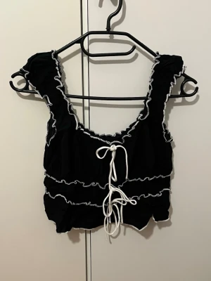 Svart croppad topp med snörning från Nelly - Säljer en svart croppad topp från Nelly i storlek XS. Toppen har vita kontrastsömmar och snörning framtill, samt volangdetaljer längs kanterna.
