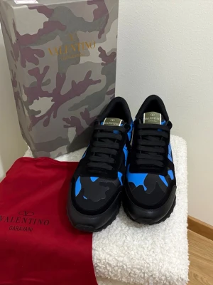 Valentino Garavani Rockrunner sneakers - Snygga Valentino Garavani Rockrunner sneakers i svart och blått med coolt kamouflagemönster. Skorna har snörning, rund tå och platt sula. Materialmix av skinn och syntet ger en lyxig känsla. Perfekta för dig som vill sticka ut med exklusiva sneakers.