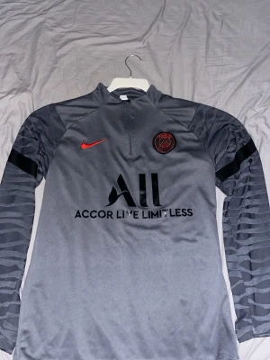 Grå Nike PSG träningsset - Säljer ett grått träningsset från Nike x PSG med långärmad tröja och matchande byxor. Tröjan har röd Nike-logga, PSG-emblem och svart tryck på bröstet samt mönstrade ärmar. Byxorna har samma röda logga och PSG-märke. Materialet är syntet och passar perfekt till fotbollsträning.