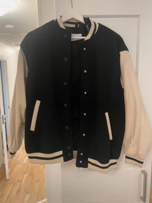 Svart och beige bomberjacka - Snygg bomberjacka i svart med beige ärmar och detaljer. Jackan har ribbade muddar med svarta och beige ränder, tryckknappar framtill och två fickor. 