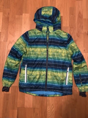 Randig skidjacka från Crivit - Snygg skidjacka från Crivit i storlek 146/152. Jackan har ett randigt mönster i blått, grönt och gult, med huva och dragkedja framtill. Två fickor med dragkedja och reflexdetalj. Perfekt för kyliga och blåsiga dagar eller utomhus aktiviteter som skid och snowboard åkning.