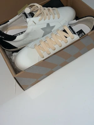 Golden Goose vita sneakers med stjärna - Golden Goose sneakers i vitt skinn med beige snörning och en grå stjärndetalj på sidan. Bakkappan är svart och glittrig med vit text. Skorna har rund tå och platt sula. Medföljer originalkartong och svart GGDB-påse.fick som jul klapp inte riktigt min stul därav säljer
