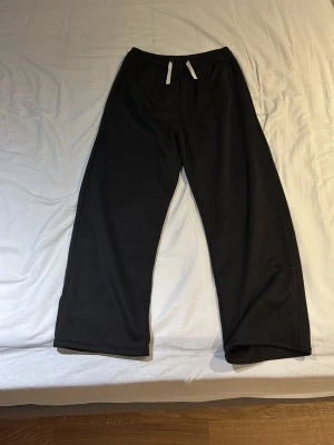 Svarta mjukisbyxor med snörning - Svarta mjukisbyxor med vit snörning i midjan och elastisk midja. Byxorna har en loose passform och raka ben, perfekta för chill dagar. Materialet känns mjukt och bekvämt mot huden. Enkla och stilrena utan synliga loggor eller mönster.