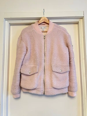 Ljusrosa teddyjacka från NLY Trend - Mysig teddyjacka i ljusrosa från NLY Trend med ribbade muddar, två stora fickor med lock och en ficka med dragkedja på ärmen. Jackan har en loose passform och stängs med dragkedja framtill. Mycket älskad och i mycket bra skick! Perfekt till vår och sommar. Säljs på flera sidor, först till kvarn. 