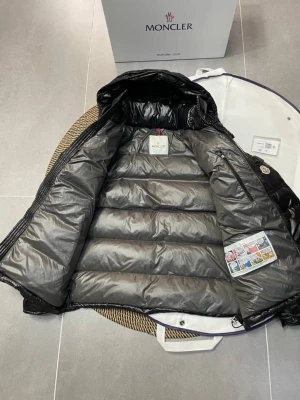 Svart Moncler dunjacka med huva - Svart dunjacka från Moncler med glansig finish och huva. Jackan har en quiltad design, dragkedja framtill och Moncler-logga på ärmen. Moncler 1: EU 36/38 (S) Moncler 2: EU 38/40 (M) Moncler 3: EU 40/42 (L)   Moncler 4: EU 42/44 (XL) Moncler 5: EU 44/46 (XXL)