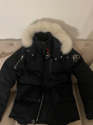 Svart pufferjacka från Moose Knuckles - Säljer en svart pufferjacka från Moose Knuckles med fluffig vit päls runt huvan. Jackan har flera dragkedjor, tryckknappar och logga på ärmen. Perfekt för kalla dagar med sin tjocka och isolerande design. Huvan är stor och ger extra värme. Den är så klart äkta.
