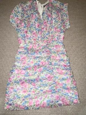 Blommig kortklänning från Zara - Säljer en söt kortklänning från Zara med färgglatt blommigt mönster i rosa, blått, lila och vitt. Klänningen har korta ärmar, v-ringad hals och är gjord i ett lätt och skönt material som passar perfekt till varmare dagar.