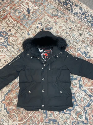 Svart pufferjacka från Moose Knuckles - Svart pufferjacka från Moose Knuckles med dunfyllning och stor huva med fluffig pälsdetalj. Jackan har dragkedja och knappar framtill, samt praktiska fickor med knappstängning. Snygga detaljer som broderad logga på insidan och dragkedja på ärmen.