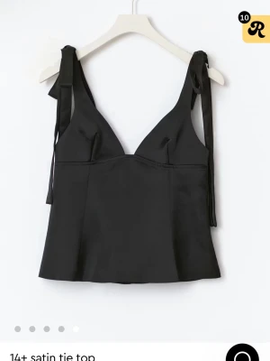 Svart satin linnetopp med knytband - Snygg svart linnetopp i satin med djup v-ringning och knytband på axlarna. Toppen är ärmlös och har en croppad passform som ger en modern och stilren look. Perfekt att matcha med jeans eller kjol för en trendig outfit. Helt oanvänd med lapp kvar, nypris 350kr, säljer för 275kr