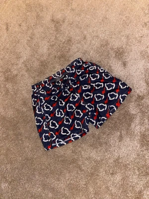 Mönstrade badshorts från Moncler - Säljer ett par snygga badshorts från Moncler i marinblått med vitt och rött mönster. Shortsen har snörning i midjan och är gjorda i polyester med meshfoder. Perfekta för sommaren och strandhäng!