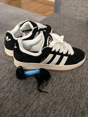 Adidas Campus sneakers svart/vit strl 42 - Säljer ett par Adidas Campus sneakers i svart mocka med vita detaljer och klassiska tre ränder på sidan. Skorna har vit platt sula, rund tå och snörning med vita skosnören. Extra svarta skosnören medföljer. Perfekt för dig som gillar retrostil och streetwear.