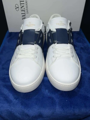 Valentino Garavani Open sneakers vit/blå - Snygga Valentino Garavani Open sneakers i vitt skinn med bred marinblå läderdetalj över mitten och vita snören. Rund tå och platt sula med mönstrad undersida. Ikonisk logga på plösen och coola nitar på sidorna. Perfekt för dig som gillar exklusiv streetstyle.