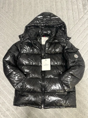 Svart pufferjacka från Moncler - Säljer en svart pufferjacka från Moncler med glansig finish och huva. Jackan har dragkedja framtill, sidofickor med dragkedja och Moncler-logga på ärmen. Fylld med dun och certifierad enligt DIST. Perfekt för kalla dagar och har en cool, modern look.