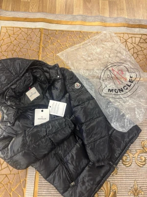 Svart pufferjacka från Moncler - Svart pufferjacka från Moncler med glansig finish och klassisk logga på ärmen. Jackan har dragkedja framtill, två sidofickor med dragkedja och snygga detaljer som tryckknappar vid kragen. Perfekt för kalla dagar och har en cool, urban vibe. Perfekt Julklapp 🎄🎅Storlek Medium