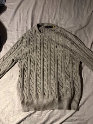 Grå stickad tröja från Polo Ralph Lauren - Säljer en snygg grå kabelstickad tröja från Polo Ralph Lauren med rund hals och diskret logga på bröstet. Tröjan är tillverkad i bomull och har ribbade muddar vid ärmslut och nederkant. Perfekt för dig som gillar klassisk och stilren design. Garanterad autenticitet. Bara prövats annars helt ny