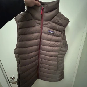 Brun dunväst från Patagonia - Säljer en brun dunväst från Patagonia i storlek M. Västen har ett quiltat mönster och dragkedja framtill. Perfekt för lager-på-lager och outdoor. Diskret Patagonia-logga på bröstet och hög krage för extra värme.