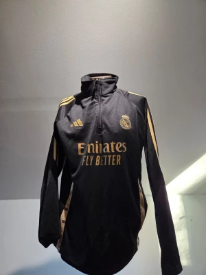 Real Madrid träningströja Adidas svart/guld - Snygg svart Real Madrid träningströja från Adidas med guldiga detaljer och loggor. Halv dragkedja framtill, långärmad och med meshpaneler på sidorna för extra ventilation. Perfekt för dig som vill ha en sportig look med stil.
