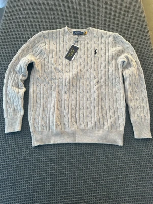 Grå stickad tröja från Polo Ralph Lauren - Snygg grå helt ny stickad tröja från Polo Ralph Lauren med klassiskt kabelstickat mönster och rund hals. Tröjan har långärmade ribbade muddar och en broderad svart logga på bröstet. Perfekt för dig som gillar stilrena och tidlösa plagg. Tröjan är i storleken L men passar mer som M