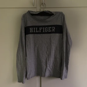 Grå långärmad T-shirt Tommy Hilfiger - Tja, säljer nu  en grå långärmad t-shirt från Tommy Hilfiger i ekologisk bomull. Den är i storlek 176, men jag skulle nog mer säga att den är i storlek XS, lite mer åt S håller. Tröjan har ett snyggt och stilrent HILFIGER-tryck över bröstet och små broderade logodetaljer i rött, vitt och blått på ärmen och halsen. Perfekt för dig som gillar stilrena och klassiska plagg. U-rigningen (kragen) på tröjan är möjligtvis något stor. Hör av er vid frågor😁