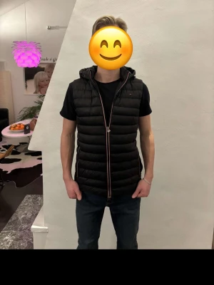 Svart dunväst från Tommy Hilfiger - Snygg svart dunväst från Tommy Hilfiger med huva och dragkedja framtill. Västen har quiltad design och diskret logga på bröstet. Perfekt att slänga över en hoodie eller t-shirt för en chill look. Modellen är 175 cm, vikt 68 kg