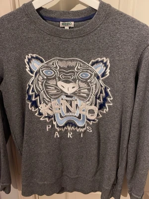 Grå sweatshirt från Kenzo med tiger - Säljer en grå sweatshirt från Kenzo med ikonisk broderad tiger i blått, vitt och silver på bröstet. Tröjan har rund hals, ribbade muddar och lång ärm. Perfekt för dig som gillar streetwear och vill sticka ut med ett coolt motiv.