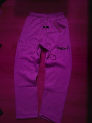 Essentials Fear of God mjukisbyxor grå - Snygga grå mjukisbyxor från Essentials Fear of God med dragsko i midjan och logga tryckt på benet. Byxorna har en relaxed fit och är tillverkade i mjukt bomullsmaterial. Perfekta för chill dagar eller streetwear-stil.