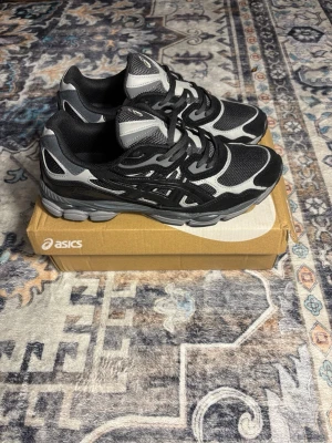 Svarta och grå Asics sneakers - Säljer ett par snygga Asics sneakers i svart och grått med coola detaljer och meshpartier för extra ventilation. Skorna har snörning och en rund tå, samt en platt sula som ger bra komfort. Perfekta för dig som gillar sportig stil och vill ha något som sticker ut.