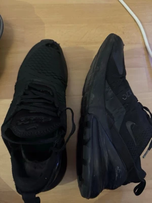 Nike Air Max 270 svarta sneakers - Svarta Nike Air Max 270 sneakers med mesh-överdel och synlig Air Max-dämpning i hälen. Skorna har snörning, platt sula och rund tå. Snygg sportig look med diskreta Nike-loggor på sidan och bak. Perfekt för dig som gillar streetwear och sportig stil.