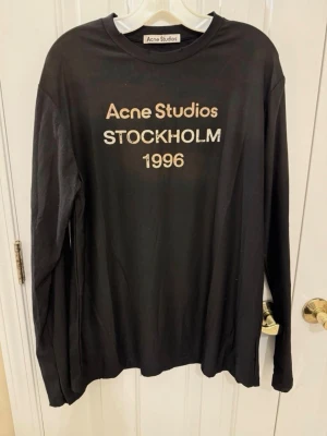 Svart långärmad t-shirt Acne Studios - Svart långärmad t-shirt från Acne Studios med tryck 'Acne Studios Stockholm 1996' på bröstet. Tillverkad i 100% bomull och har en klassisk rund hals. Perfekt för dig som gillar stilrena och ikoniska plagg med streetkänsla.