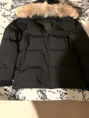Canada Goose Wyndham Parka - Säljer en svart dunjacka från Canada Goose i storlek L med black label. Jackan är perfekt för kalla vinterdagar med sin tjocka vaddering och avtagbar pälskrage. Den har en dragkedja och knappar framtill samt en broderad logga på ärmen. Jackan är i nyskick och kommer med originalpåse.