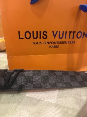 Louis Vuitton rutig skinnbälte 100cm - Säljer ett Louis Vuitton bälte med klassiskt rutigt mönster i svart och grått. Bältet har en stilren LV-spänne i svart metall och är tillverkat i äkta skinn. Insidan är ljusbrun och präglad med Louis Vuitton Paris, made in France. Perfekt accessoar för att lyfta din outfit.