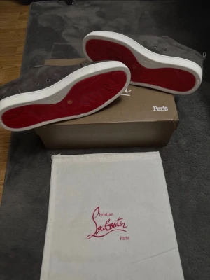 Christian Louboutin grå mocka sneakers - Snygga sneakers från Christian Louboutin i grå mocka med klassisk röd sula och vita kanter. Skorna har rund tå, snörning och beige innersida. Kommer med kartong och dustbag med Louboutin-logga. Skorna är i storlek: 42