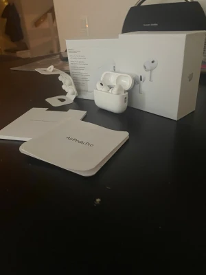 AirPods Pro 2 - Apple AirPods Pro med laddningsetui, extra öronkuddar och originalförpackning. Komplett med manual och tillbehör. Hörlurarna ser ut att vara i mycket gott skick utan synligt slitage. Perfekt för brusreducering och trådlös musikupplevelse.