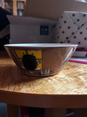 Brun Mumin-skål med stinky in action  - Säljer min Mumin-skål från Arabia med motiv av Stinky in Action. Fick en i present och har redan en så har dubbletter. Denna är alltså helt ny men lapp och allt. Skålen har en vit insida och färgglada illustrationer i svart, gul och orange på utsidan. Perfekt till frukost eller snacks!