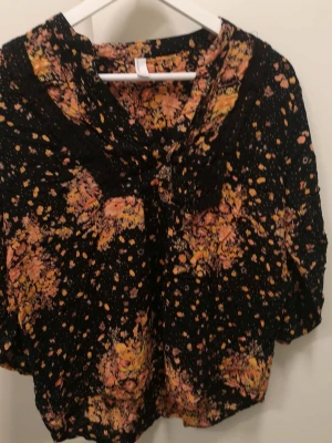 Vera Moda svart/orange blus -  Svart blus med ett blommigt mönster i orange toner. Blusen har en v-ringning med spetsdetaljer och 3/4 ärmar.