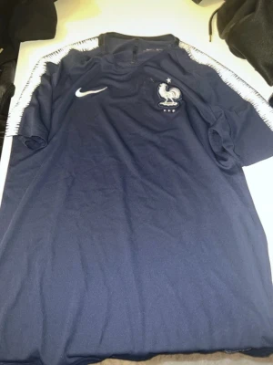 Marinblå Nike fotbollströja Frankrike - Säljer en marinblå fotbollströja från Nike med Frankrikes landslagsmärke på bröstet och vita detaljer längs axlarna. Tröjan har rund hals och är tillverkad i ett lätt syntetmaterial som andas. Perfekt för dig som älskar fotboll och vill representera Frankrike.