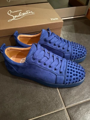 Blå Christian Louboutin sneakers - Säljer ett par blå sneakers från Christian Louboutin med coola nitar på tån och klassisk röd sula. Skorna är gjorda i mocka med snörning och har en rund tå. Insidan är beige och har guldfärgad logga. Perfekt för dig som vill sticka ut med din stil. Aldrig använda. Kvitto saknas.