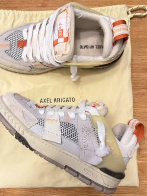 Axel Arigato sneakers i vitt och orange - Säljer ett par feta Axel Arigato sneakers i storlek 42. Skorna har en mix av vitt, beige och orange med coola meshpaneler, mockadetaljer och kontrastsömmar. Platt sula, rund tå och snörning. Märket syns på sidan och tungan. Perfekt för dig som gillar streetstyle.