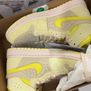 Nike Jordan 1 Low i gul och beige - Snygga Nike Jordan 1 Low sneakers i en fräsch kombination av ljusgul mocka och beige detaljer, med en tydlig gul swoosh i textil. Skorna har klassisk snörning och platt sula, perfekt för dig som vill sticka ut med färg. Kommer i originalkartong.