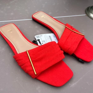 Röda sandaler från Zara med gulddetalj - Snygga röda sandaler från Zara med fyrkantig tå och bred rem framtill. Remmen har en elegant guldfärgad metallstav som detalj. Skorna är tillverkade i ett tygmaterial och har platt sula, perfekt för en trendig look.🤩Aldrig använda och prislappen är kvar. 