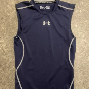 Mörkblå träningstopp från Under Armour - Säljer en mörkblå ärmlös träningstopp från Under Armour med silverfärgad logga på bröstet och kontrastsömmar. Tight passform och tillverkad i syntetmaterial som andas, perfekt för intensiva träningspass.