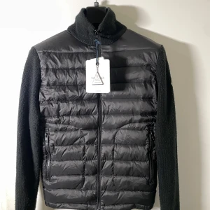 Svart dunväst från Moncler med stickade ärmar - Svart dunväst från Moncler med quiltad framsida och stickade ärmar. Västen har hög krage, dragkedja framtill och två fickor med dragkedja. Moncler-logga på vänster ärm. Perfekt för lager-på-lager och snygg till streetwear.