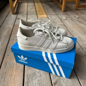 Adidas Superstar vita sneakers 37⅓ - Säljer ett par klassiska Adidas Superstar sneakers i helvitt skinn med ikoniska tre ränder på sidan och snörning. Skorna har rund tå och platt sula, perfekt för en clean och tidlös look. Levereras i originalkartong.