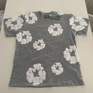 Grå t-shirt med vita blommor Denim Tears - Grå t-shirt från Denim Tears med stora vita blommiga tryck över hela plagget. Klassisk passform och rund halsringning. Tillverkad i mjuk bomull och har en unik, modern vibe. Perfekt för dig som vill sticka ut med en cool och stilren look.
