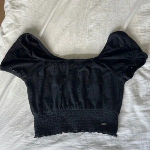 Svart croppad topp från Hollister - Svart croppad topp från Hollister med smockad midja och broderade hålmönster. Toppen har bred resår vid axlarna och kan bäras offshoulder. Tillverkad i mjuk bomull, perfekt för en trendig och avslappnad look.