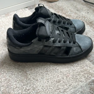 Adidas Campus sneakers i mörkgrå mocka - Säljer ett par Adidas Campus sneakers i mörkgrå och svart mocka med klassiska tre ränder på sidan. Skorna har rund tå, platt sula och svart snörning. Adidas-logga på hälen och tungan. Perfekt för dig som gillar stilrena och tidlösa sneakers.