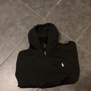 Svart hoodie från Polo Ralph Lauren - Svart hoodie från Polo Ralph Lauren med dragkedja och klassisk vit broderad logga på bröstet. Tröjan har huva med snörning och långa ärmar. Perfekt för en chill och stilren look.
